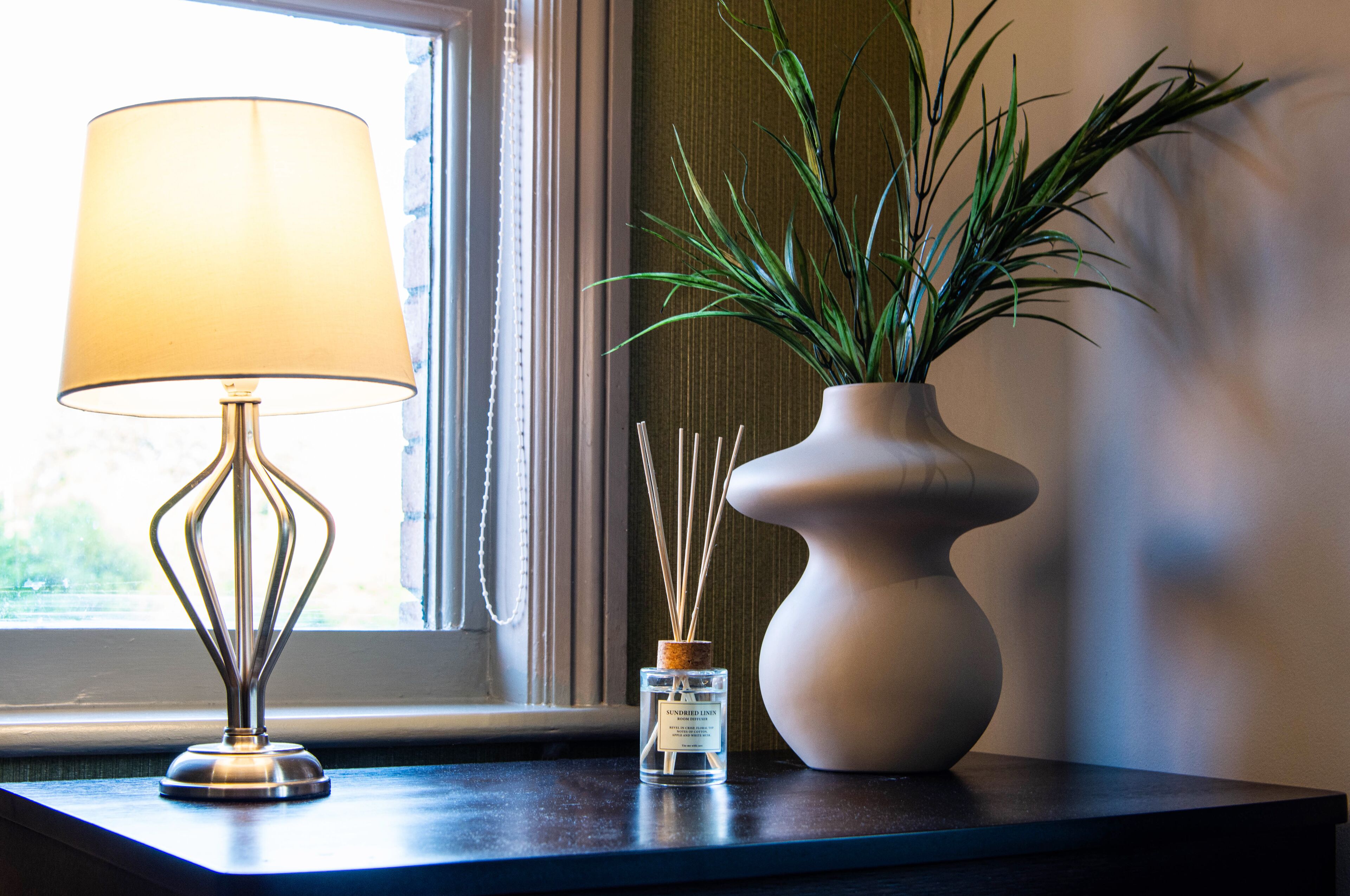 A table with a lamp, a small bottle of reed diffuser, and a vase with a plant.