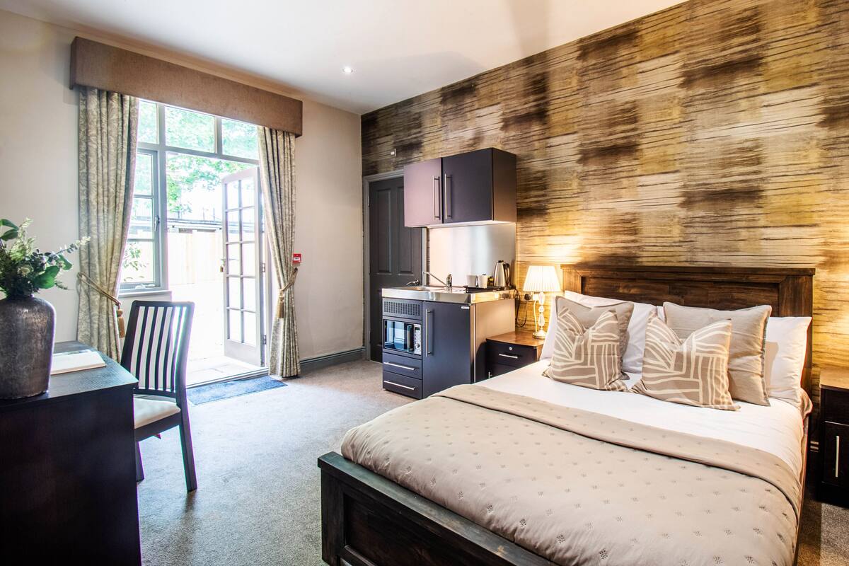 A hotel room with a bed, bedside tables, a desk, a chair, a kitchenette, and a large window with curtains.