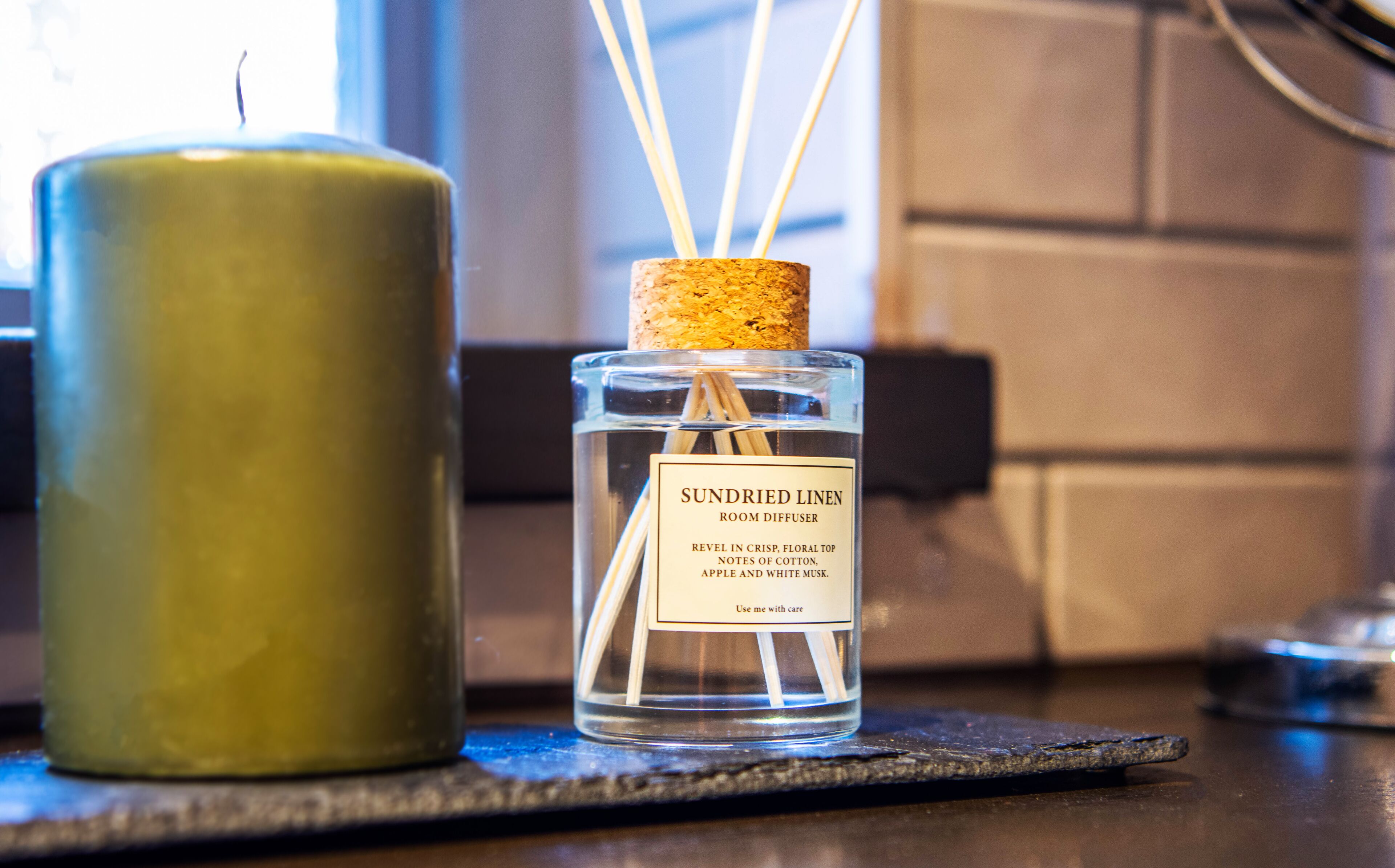 A room diffuser with a cork stopper and reed sticks, next to a green candle on a dark surface.