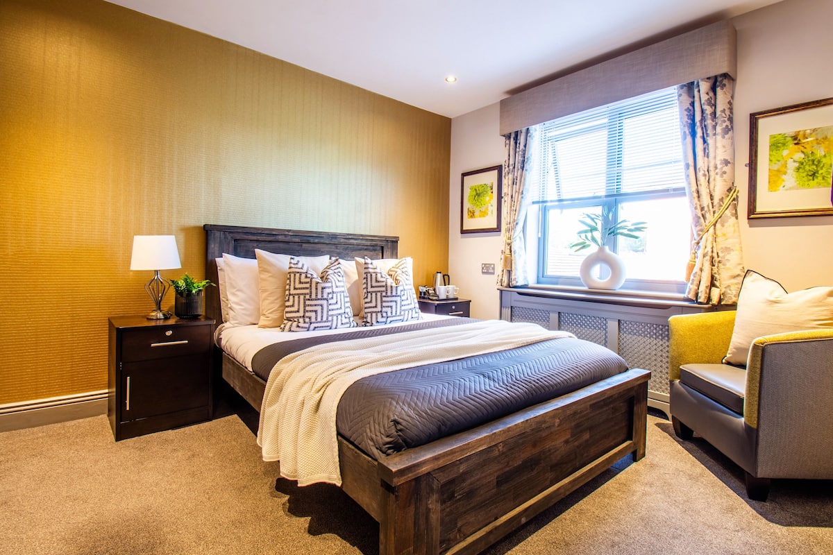 A neatly arranged bedroom with a large bed, a bedside table with a lamp, and a window with curtains.