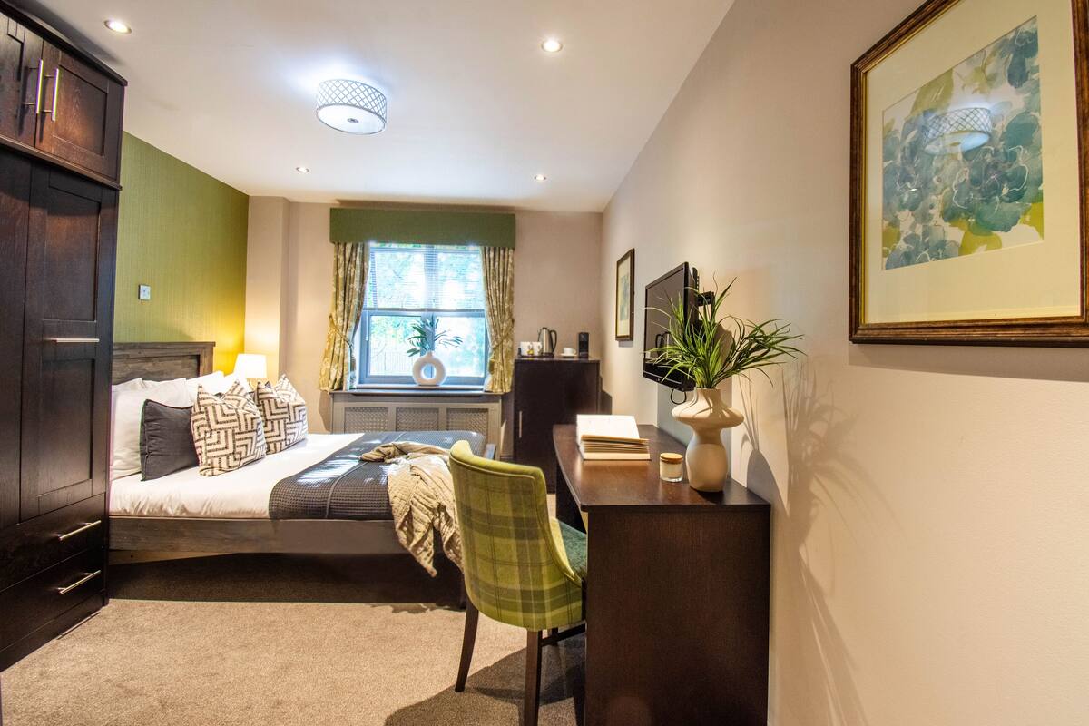 A bedroom with a bed, bedside table, chair, window with curtains, and a painting on the wall.