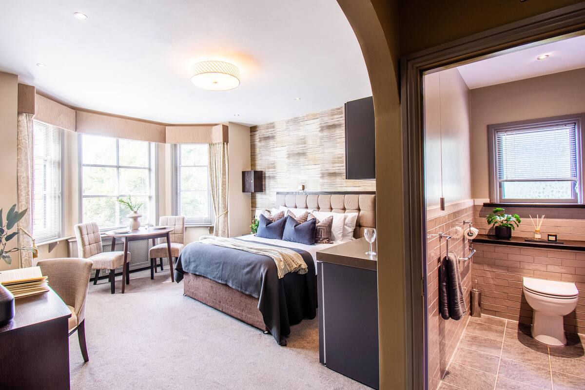 A modern hotel room with a large bed, a sitting area with a table and chairs, a small desk with a vase, and a view of the outside through large windows.