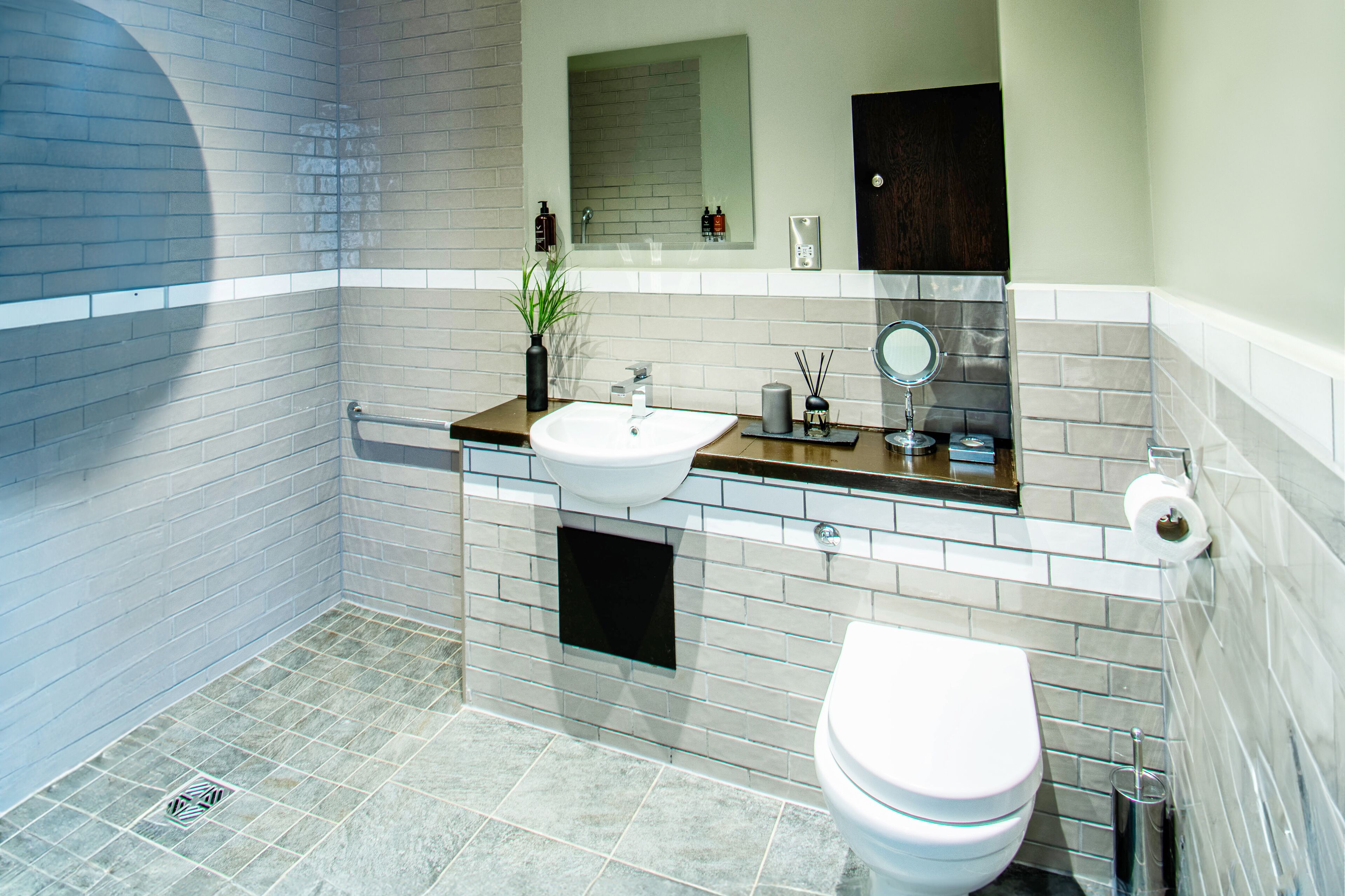 A modern bathroom with a white sink, mirror, and toilet, tiled walls, and a small plant on the counter.