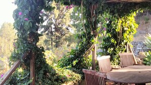 Zona relax esterna in legno con tavolo e sedie, immersa nel verde e delimitata da un muro in pietra.