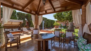 Een terras buiten met een houten tafel en stoelen, een overdekte zithoek, en uitzicht op de tuin.