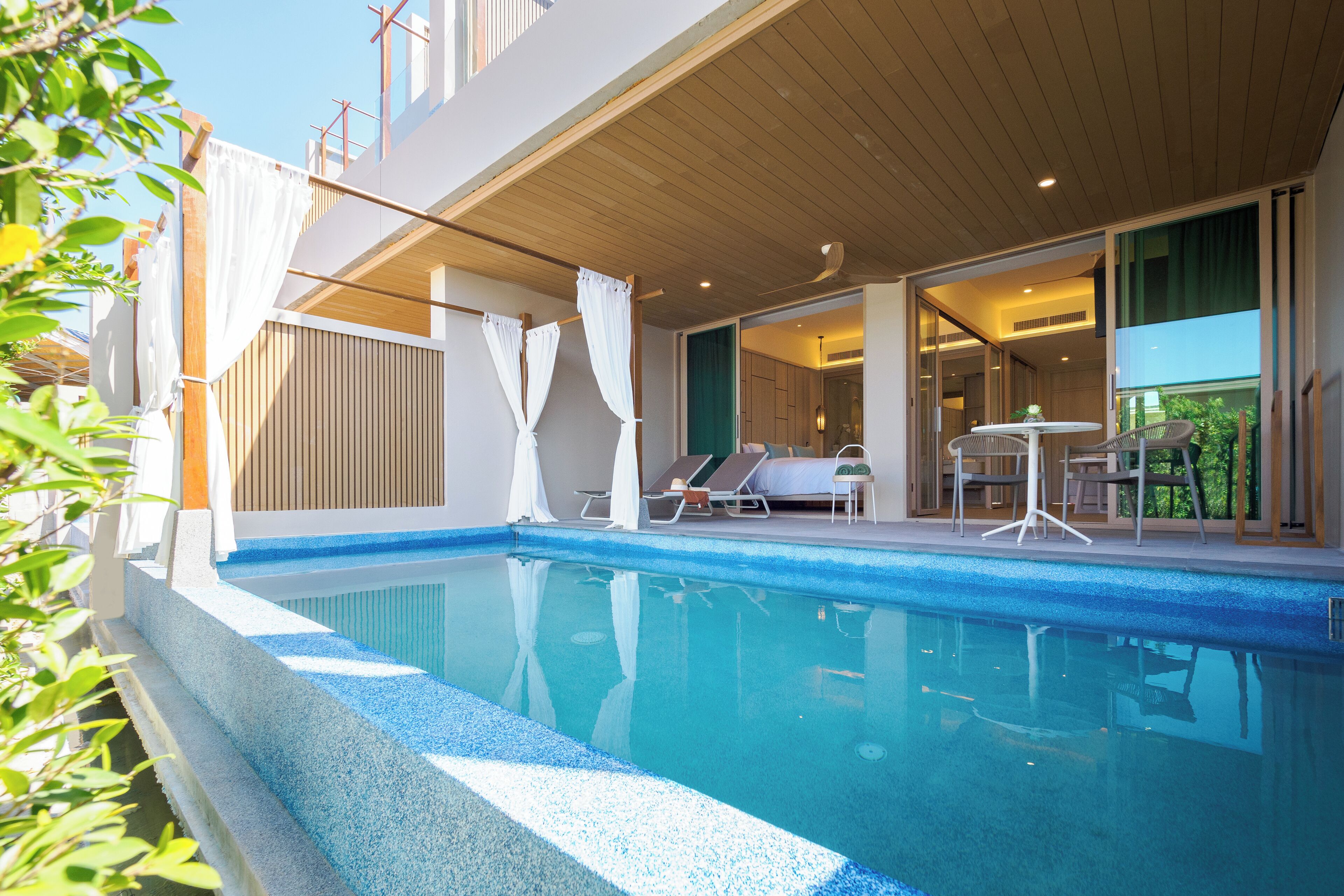 A swimming pool with lounge chairs and a table, a covered outdoor area, and a view of the surrounding greenery.