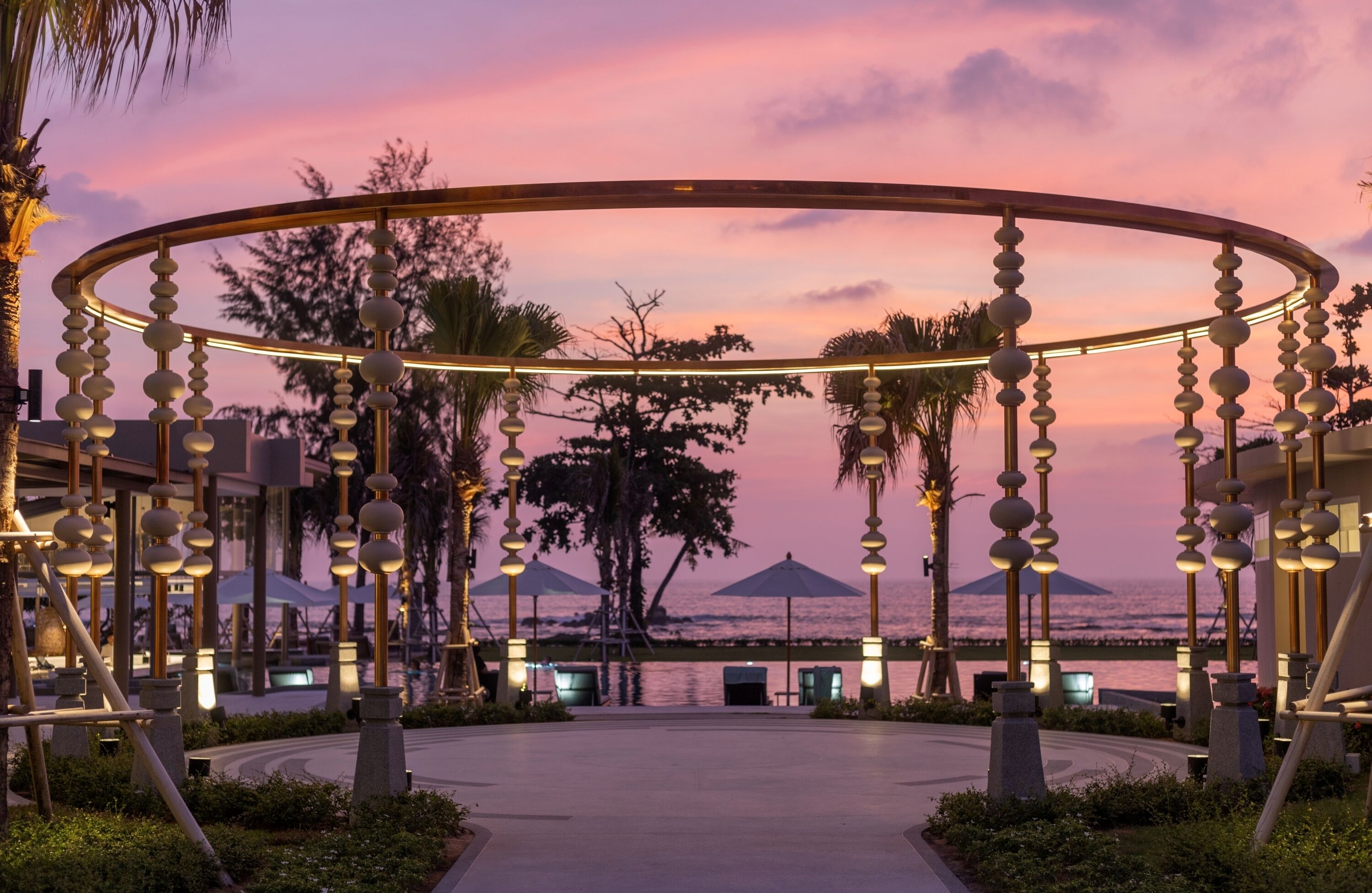 A circular outdoor area with a railing, palm trees, and a view of the sea at sunset.