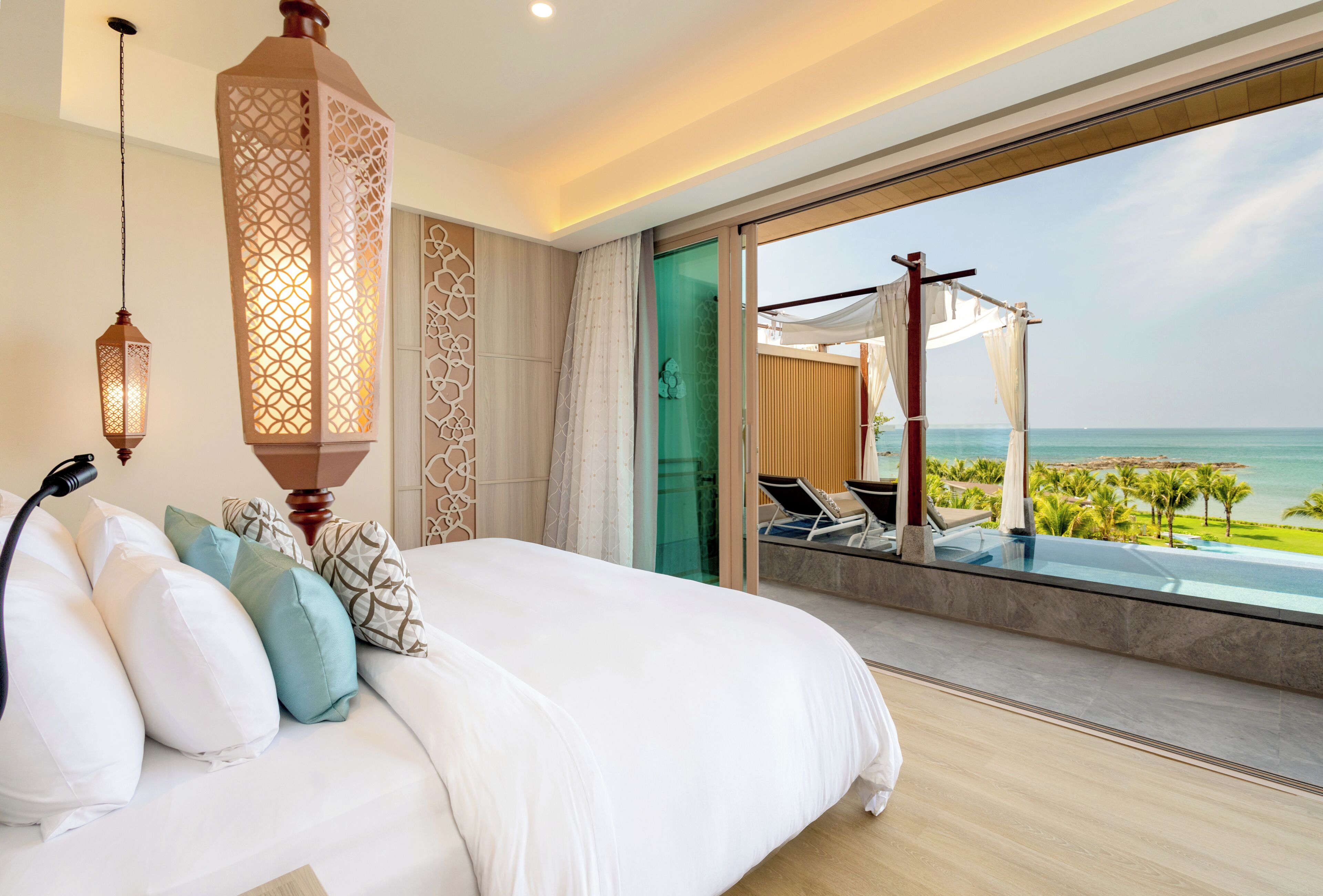 A bedroom with a large bed, a view of the ocean, and a balcony with lounge chairs.