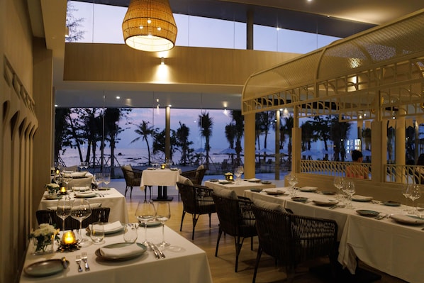 A restaurant with tables set for dining, overlooking a beach with palm trees.