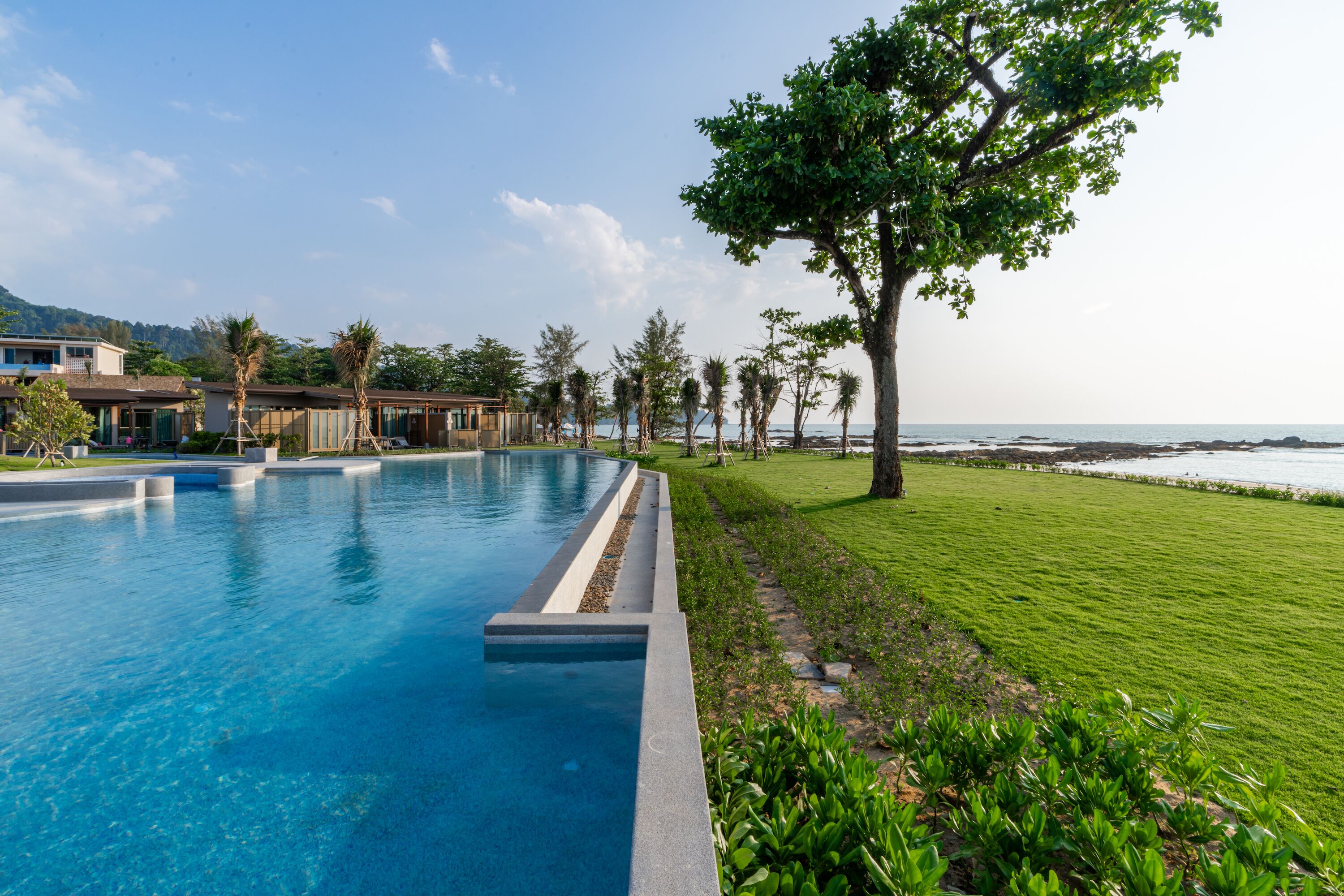 A swimming pool with a view of the ocean, surrounded by a well-maintained lawn and trees.