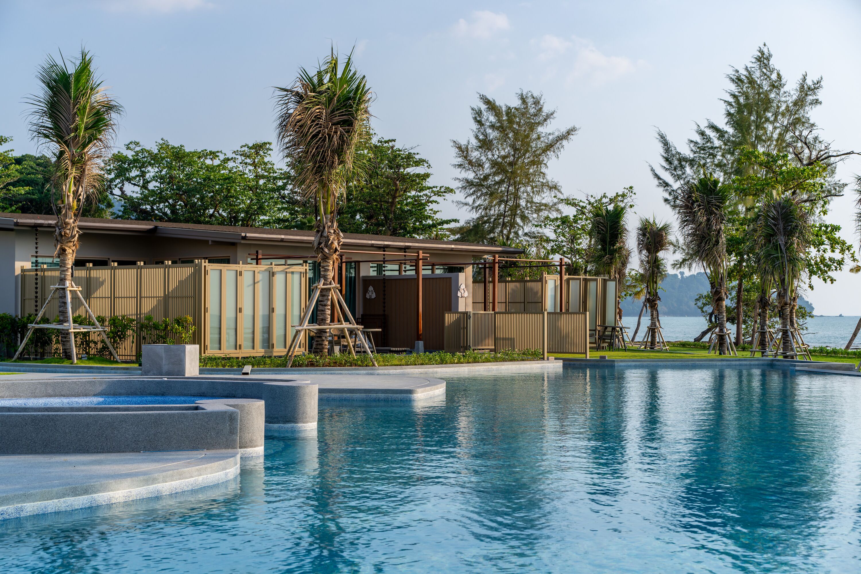 A pool area with a modern building, palm trees, and a view of the sea.