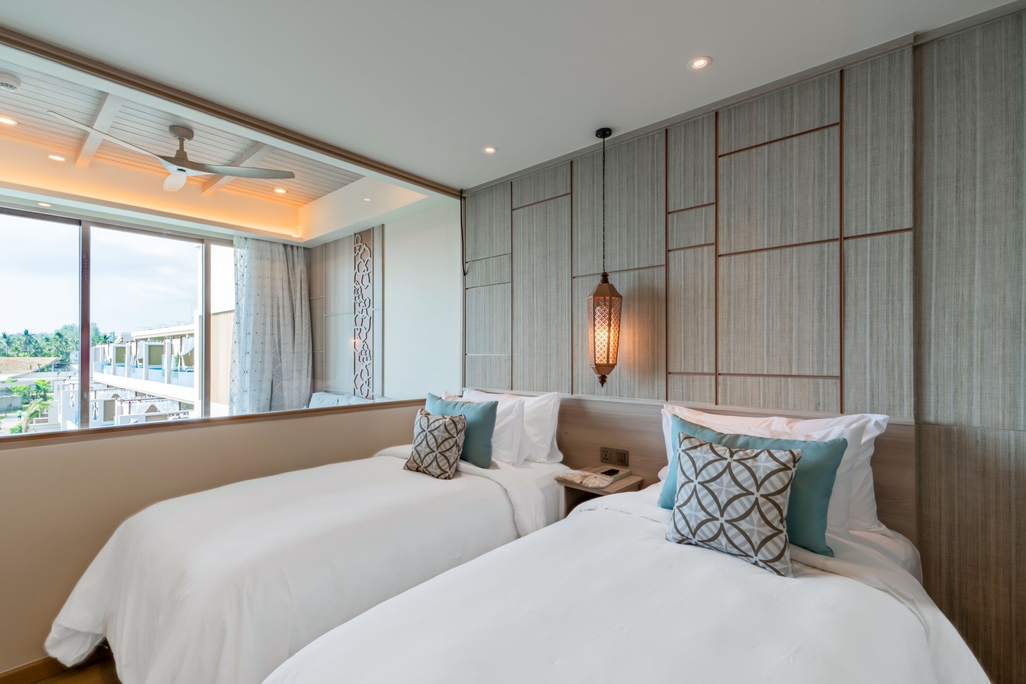 Une chambre d’hôtel avec deux lits, un ventilateur de plafond, une grande fenêtre donnant sur un beau paysage et un luminaire fixé au mur.