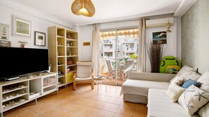 A living room with a television, a sofa, a bookshelf, and a balcony with a view of buildings. - Vacation Apartment 'La Casita De La Tejita' with Shared Pool and Wi-Fi (Granadilla)