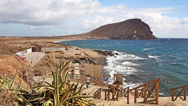 A coastal beach with a rocky hill, wooden stairs, and a sandy area with people. - Vacation Apartment 'La Casita De La Tejita' with Shared Pool and Wi-Fi (Granadilla)