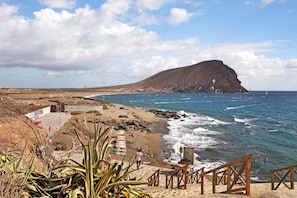 A coastal beach with a rocky hill, wooden stairs, and a sandy area with people.