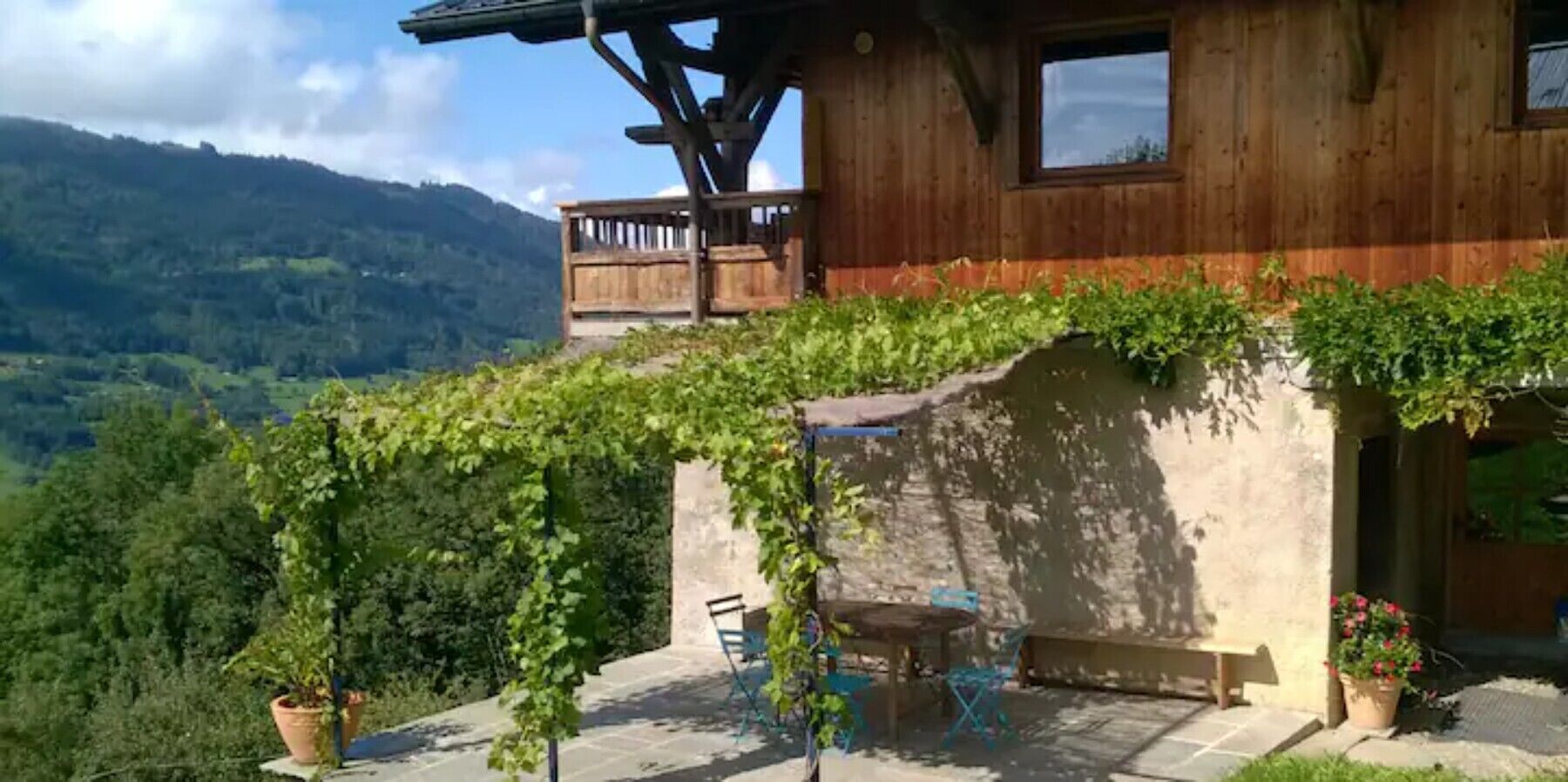 Una baita in legno con un balcone, un tavolo e sedie all'esterno, circondata da vegetazione e montagne.