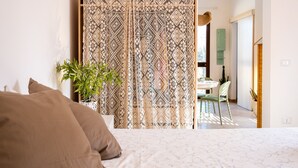 Ein Schlafzimmer mit einem Bett, einem gemusterten Vorhang, einer Topfpflanze und Blick in einen Essbereich.