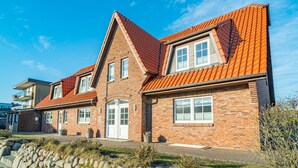 A two-story brick house with a red-tiled roof, a stone wall in the foreground, and a clear blue sky. - Appartement Dünenglück (Wenningstedt-Braderup (Sylt))