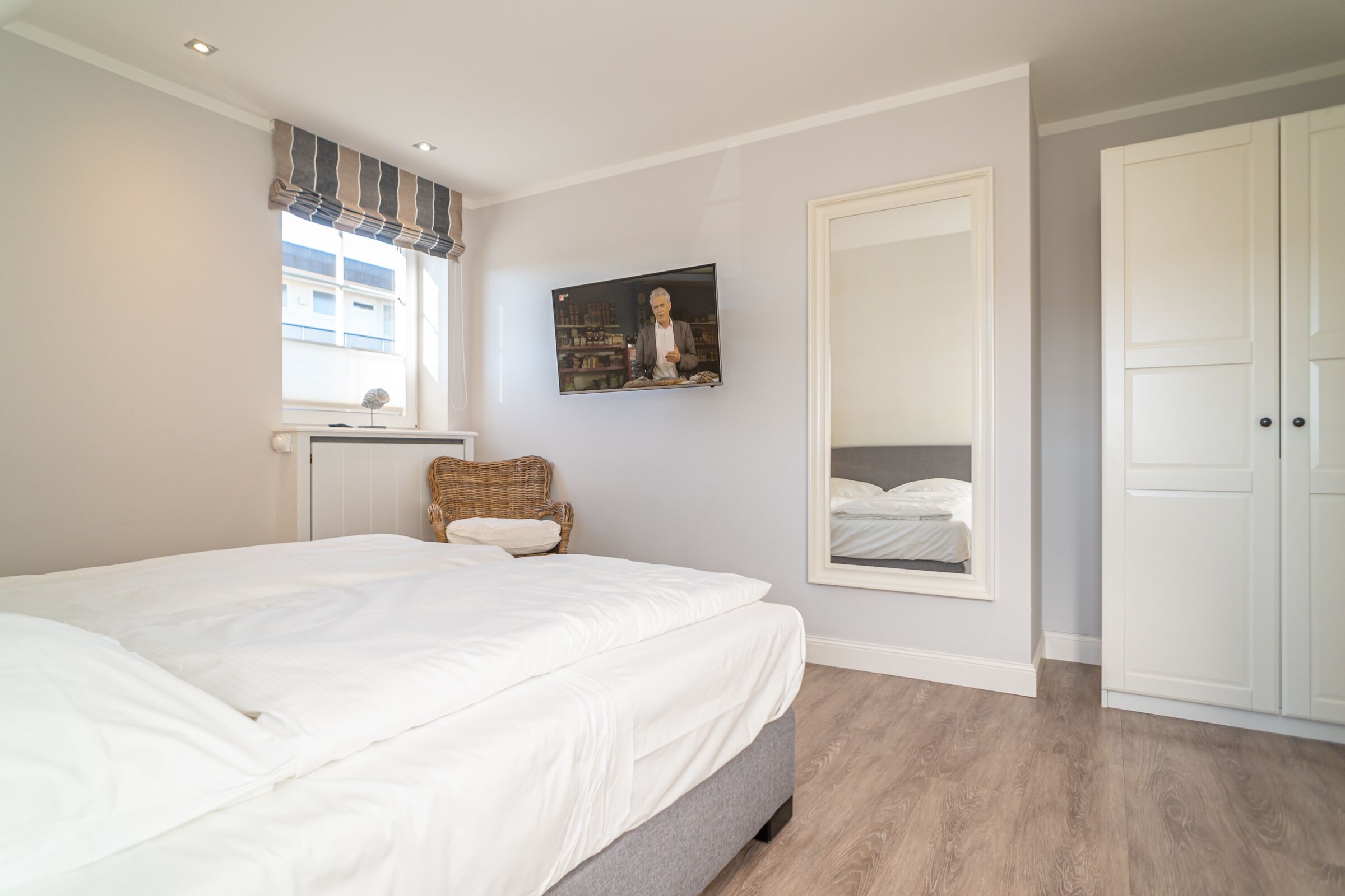 Een slaapkamer met een groot bed, een rieten stoel, een raam met gordijnen, een wandtelevisie en een kledingkast.