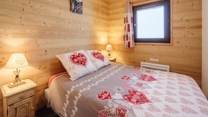 A wooden cabin bedroom with a bed, nightstand, lamp, and a window with curtains. - Apartment 'Le Petit Bonheur' with Mountain View and Balcony (Modane)