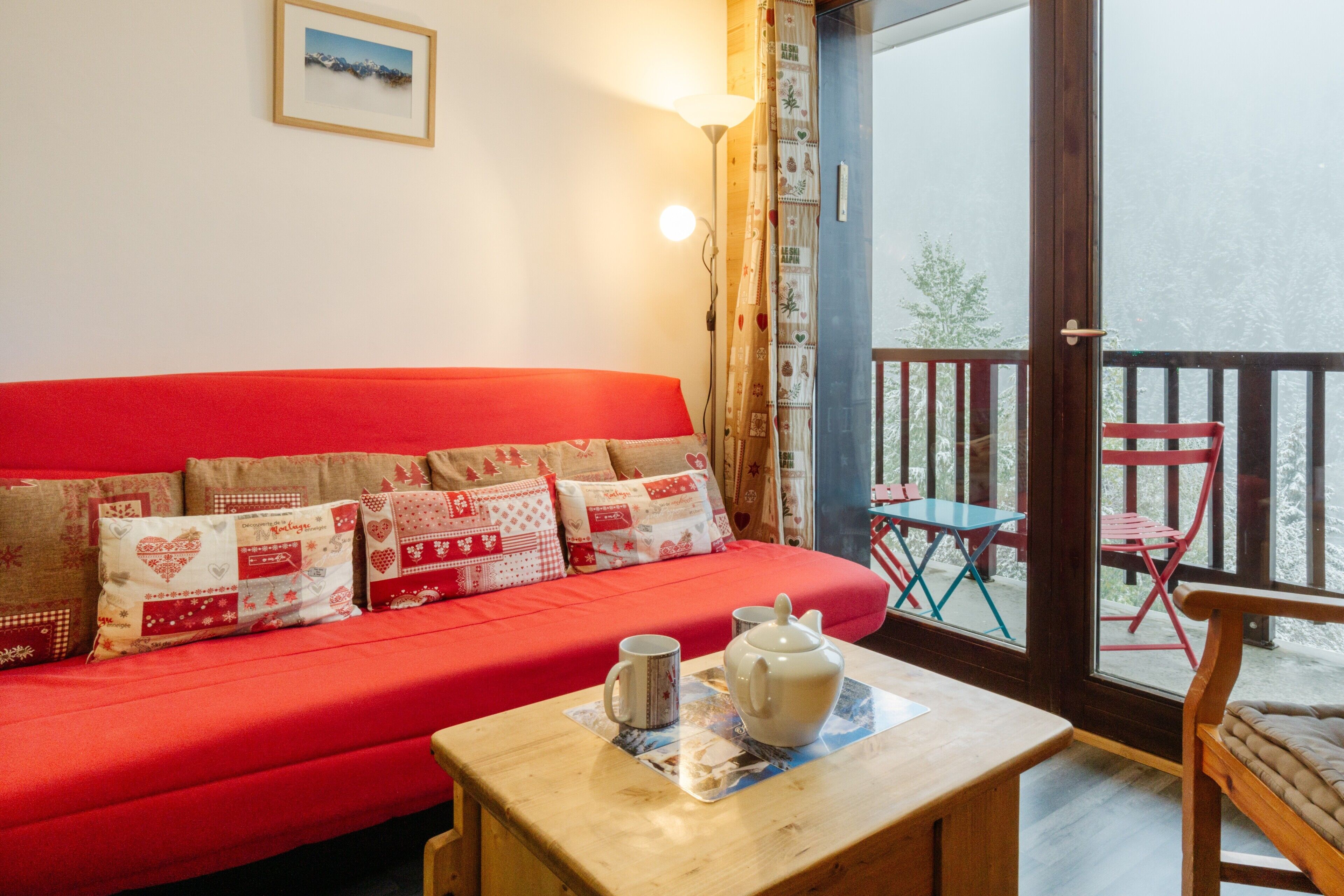 Ein rotes Sofa mit gemusterten Kissen, ein Holztisch mit Teekanne und Tasse, und Blick auf Sitzgelegenheiten im Freien durch eine Glastür.