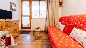 Un salon chaleureux avec un meuble TV en bois, un canapé rouge agrémenté de coussins à motifs, une petite table ronde sur laquelle se trouve une tasse, et une fenêtre avec des rideaux.