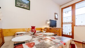 Un coin repas avec une table dressée pour deux personnes, un téléviseur posé sur un meuble et une fenêtre donnant sur un paysage montagneux.