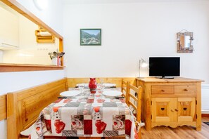 Uma área de refeições com mesa de madeira posta para quatro pessoas, uma televisão em um móvel de madeira e um quadro na parede.