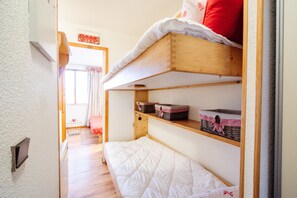 Um corredor estreito com uma cama de solteiro de madeira, uma mesinha e cestos em prateleiras.