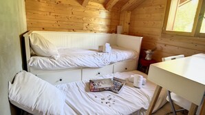 Een kleine, knusse kamer met houten wanden en een schuine kap. Er staat een stapelbed met wit beddengoed en een klein tafeltje met een doos chocolade en een beker.