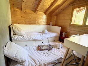 A small, cozy room with wooden walls and a sloped ceiling. There is a bunk bed with white bedding and a small table with a box of chocolates and a cup.