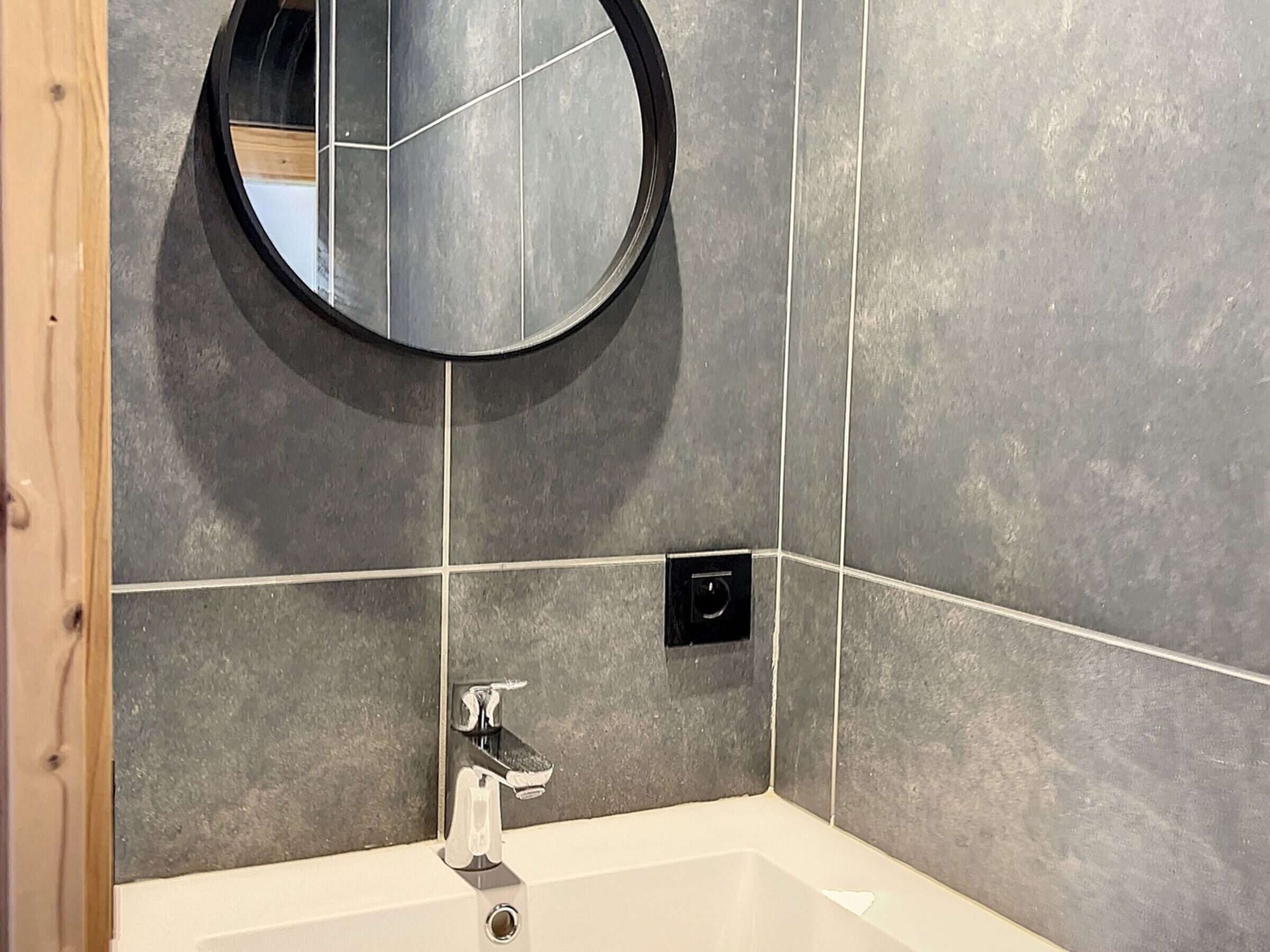 Un baño con lavamanos blanco, un espejo redondo y paredes de azulejos grises.