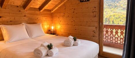Dormitorio de cabaña de madera con cama, almohadas, mesita de noche con lámpara y vista a un bosque desde un balcón.