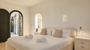 Een slaapkamer met een groot bed, een nachtkastje, een lamp en uitzicht door een boogvormige deuropening.