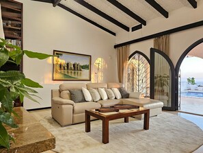 Una sala de estar con un sofá color beige, una mesa de centro de madera, un cuadro y vistas a una piscina a través de una ventana en arco.