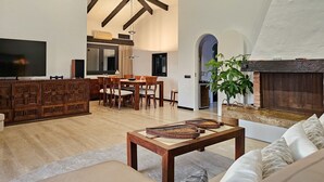 Een ruime woonkamer met een hoge plafond, houten balken, een open haard, een eethoek en een televisie.