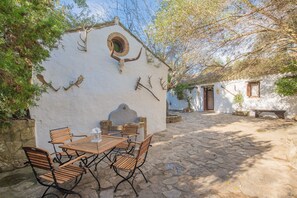Un espacio de comedor al aire libre de estilo rústico, con patio de baldosas de piedra, mesa y sillas de madera, y un edificio blanco con motivos decorativos de astas.