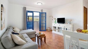 A living room with a sofa, a TV, a dining table with a basket of fruit, and a white cabinet. - Holiday Home 'Casa Famara' with Sea Views, Private Terrace and Wi-Fi (Caleta de Famara)