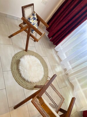 Quarto com duas cadeiras de madeira com encosto trançado, um tapete redondo trançado e um travesseiro branco fofo, além de uma cortina vermelha parcialmente aberta.