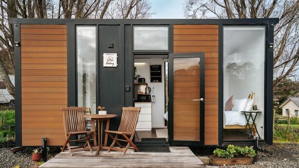 A compact, modern cabin with a wooden exterior and a glass door leading to a kitchenette. Inside, there is a bed with a nightstand and a small table.