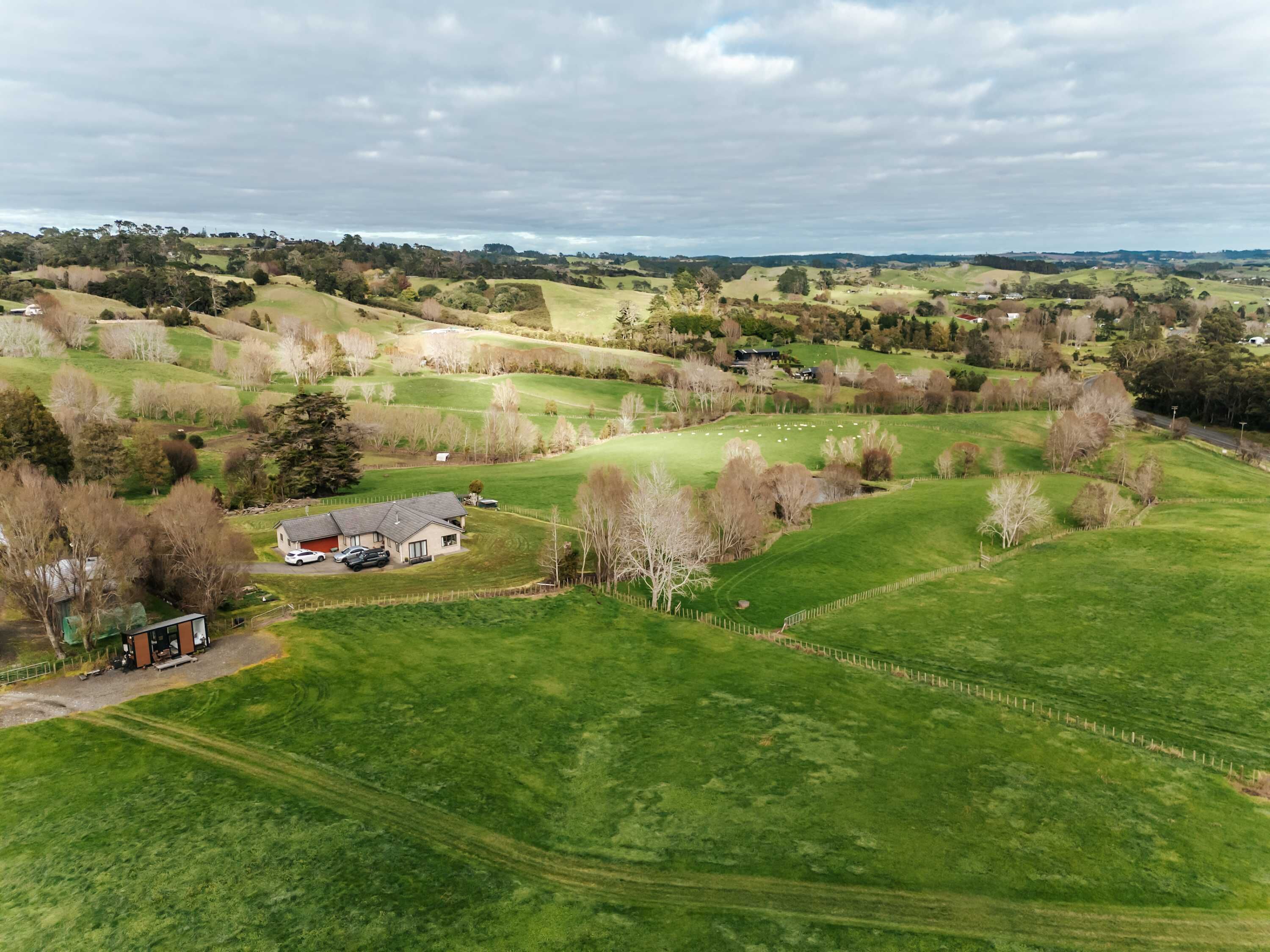 Ländliche Landschaft mit sanften Hügeln, einem eingezäunten Feld und einem kleinen Haus mit geparkten Autos.
