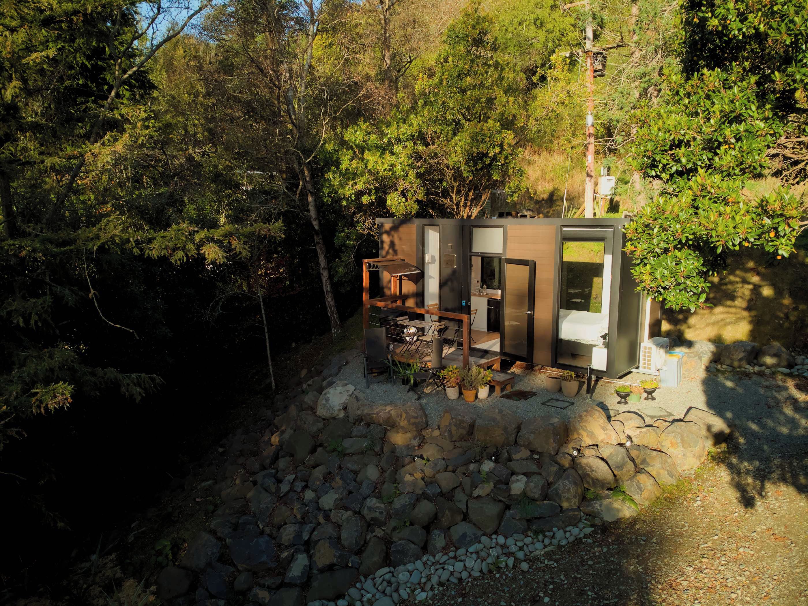 A modern cabin with a stone foundation, surrounded by dense forest. It features a compact layout with a dining area and a small kitchen.