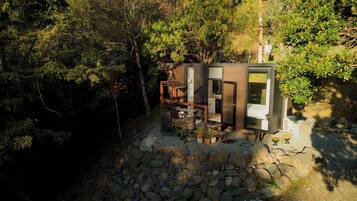 A modern cabin with a stone foundation, surrounded by dense forest. It features a compact layout with a dining area and a small kitchen.