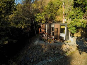 A modern cabin with a stone foundation, surrounded by dense forest. It features a compact layout with a dining area and a small kitchen.
