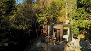A modern cabin with a stone foundation, surrounded by dense forest. It features a compact layout with a dining area and a small kitchen. - Cosy Dell Tiny House (Herbert)