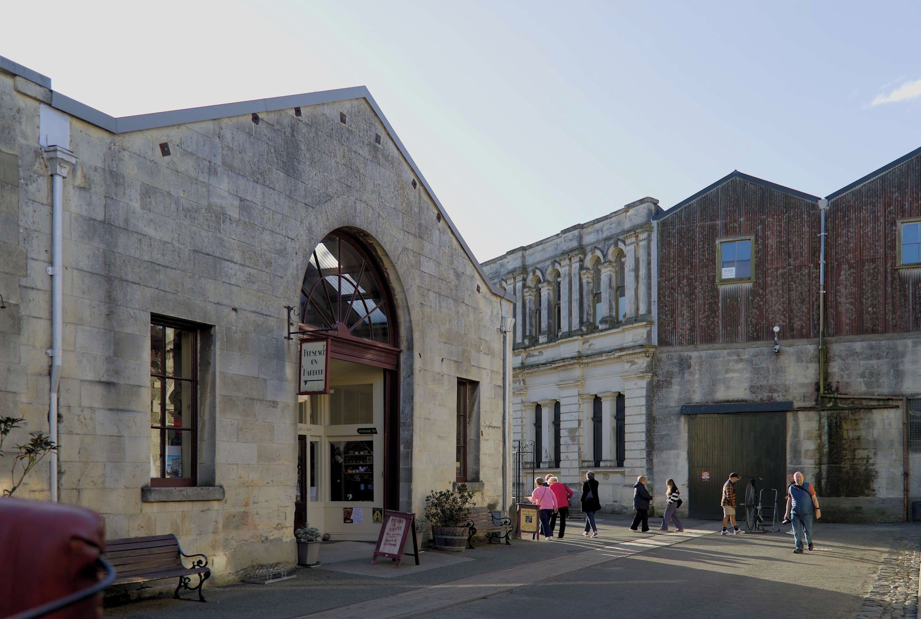 A stone building with a large arched entrance, a bench outside, and a few people walking by.