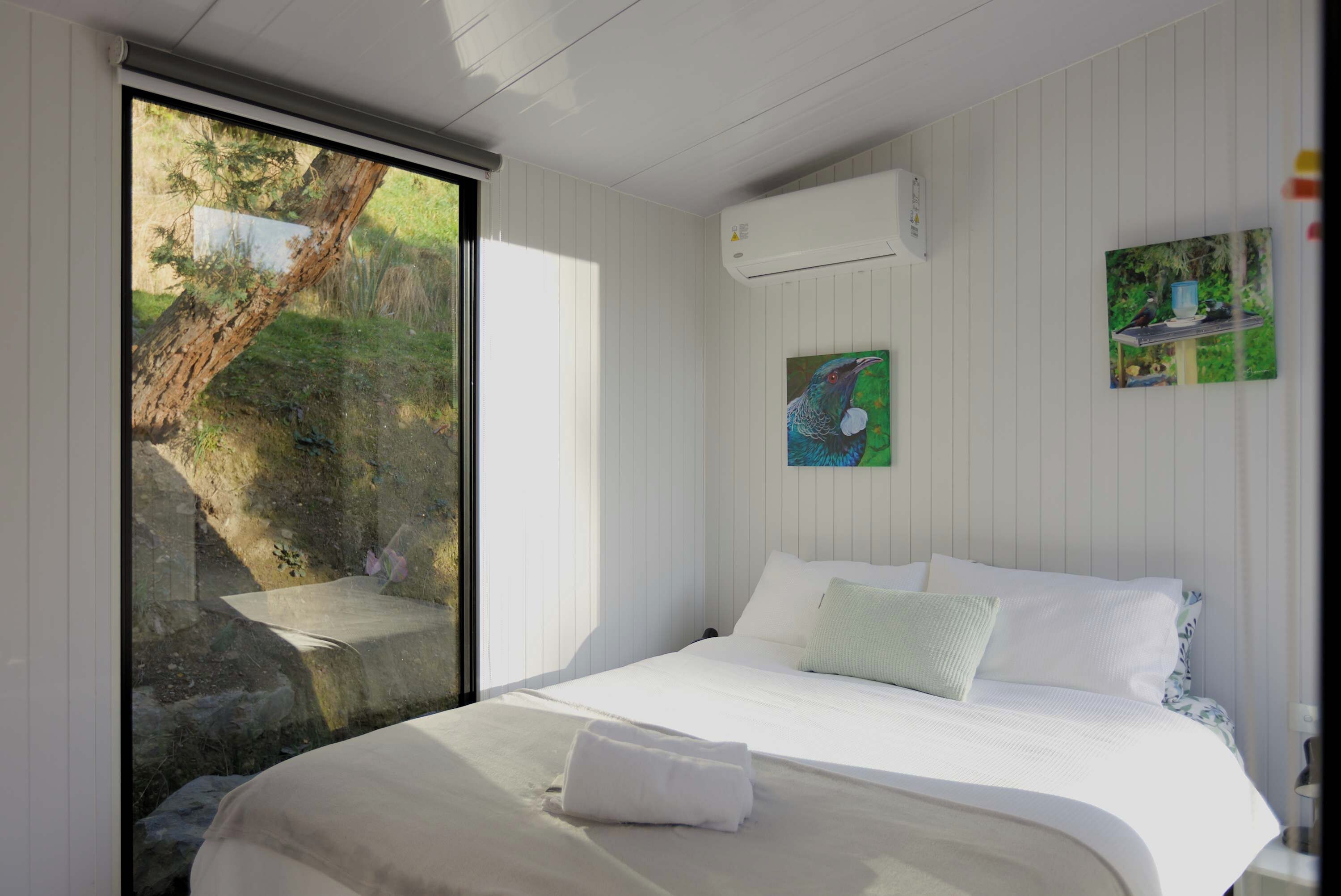 Une chambre avec une grande fenêtre, un lit recouvert de draps blancs et un tableau accroché au mur.