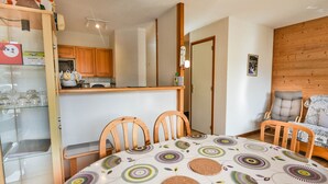 A dining area with a round table covered with a patterned tablecloth, two wooden chairs, a wooden chair with a cushion, a kitchen area with wooden cabinets, and a wooden wall panel. - Les Gets - 2BR Apartment for 6 with Balcony and WIFI (Les Gets)