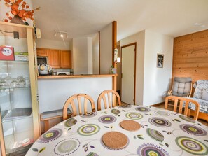 Ein Essbereich mit einem runden Tisch, bedeckt mit einer gemusterten Tischdecke, zwei Holzsesseln, ein Holzstuhl mit Kissen, eine Küchenzeile mit Holzschränken und einer Holzwandverkleidung.