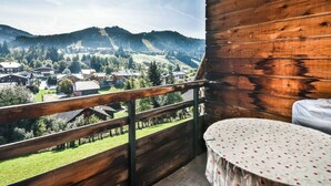 A balcony with a wooden railing, a round table with a floral tablecloth, and a view of a village and mountains in the background. - Les Gets - 2BR Apartment for 6 with Balcony and WIFI (Les Gets)